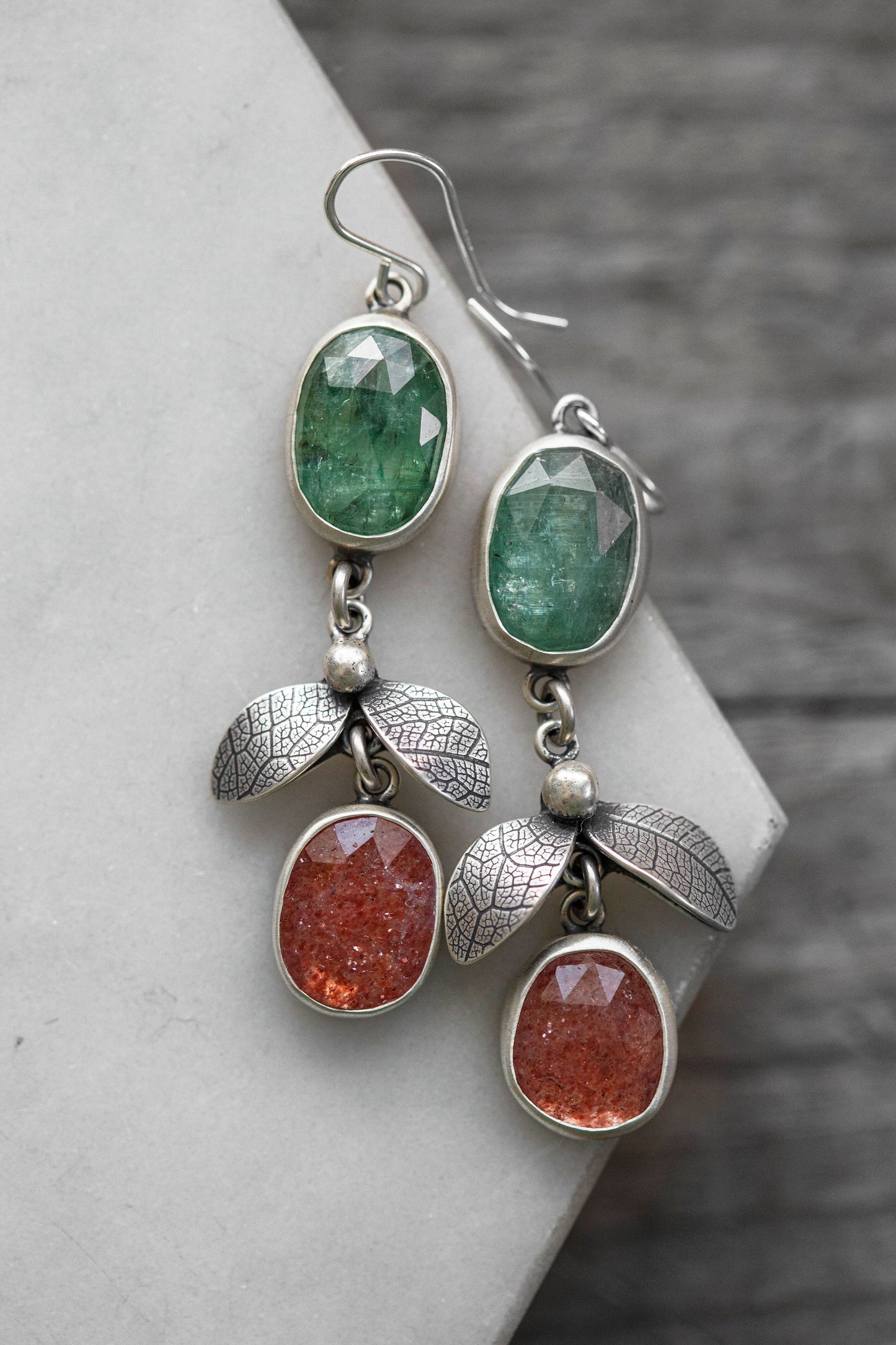 A pair of silver dangle earrings with green faceted stones at the top and textured silver leaf charms between oval orange-red stones, displayed on a light gray stone slab against a dark wood background.