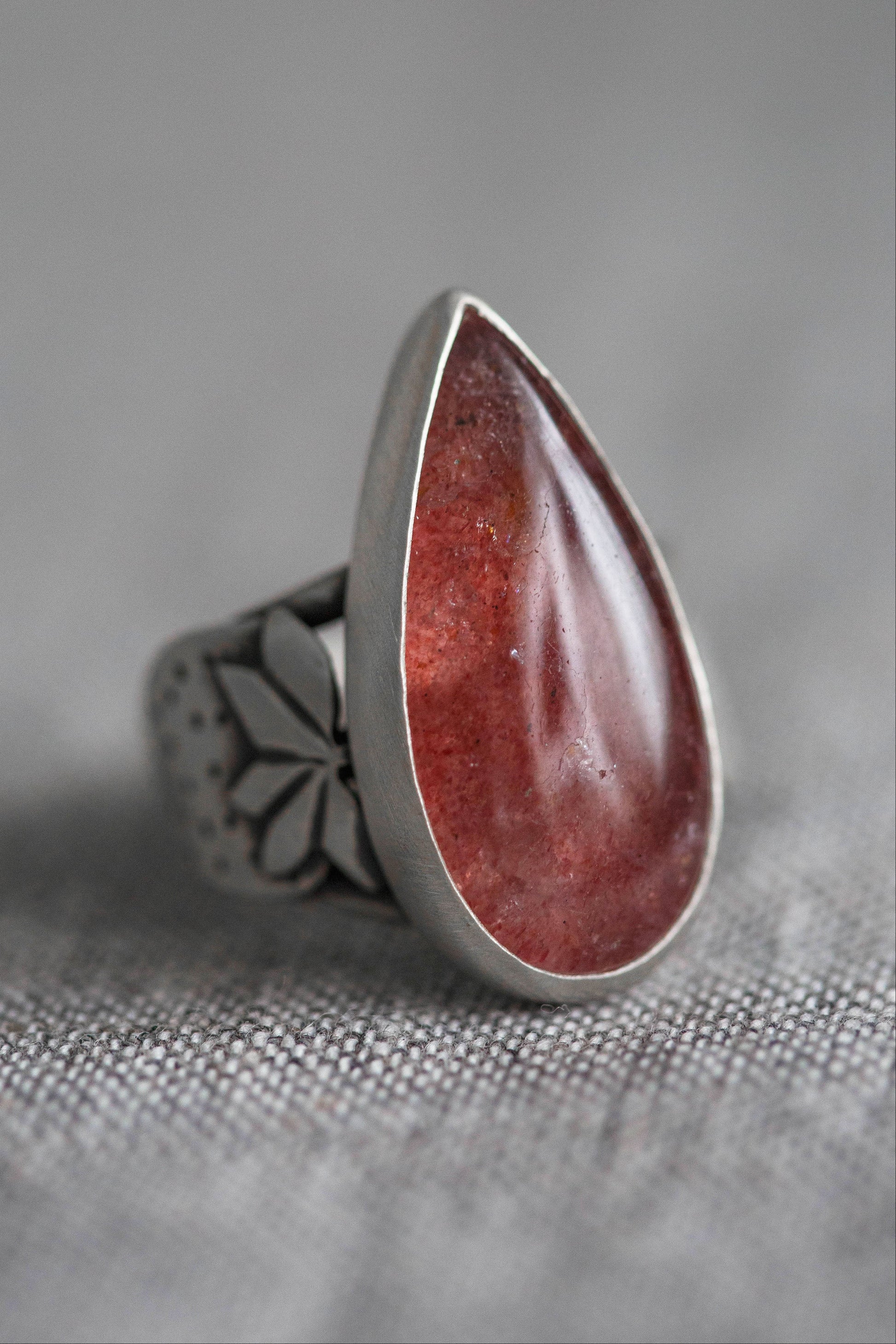 A silver ring featuring a large, teardrop-shaped red gemstone, photographed close-up on a textured gray fabric background.