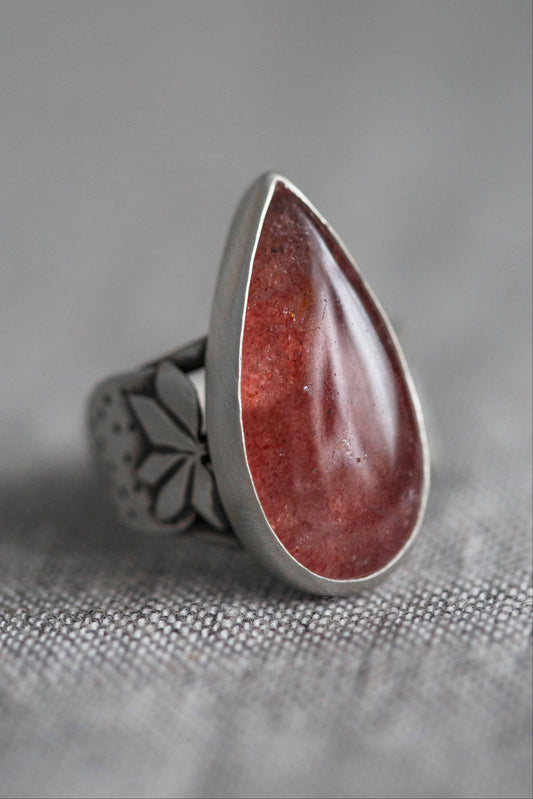 A silver ring featuring a large, teardrop-shaped red gemstone, photographed close-up on a textured gray fabric background.