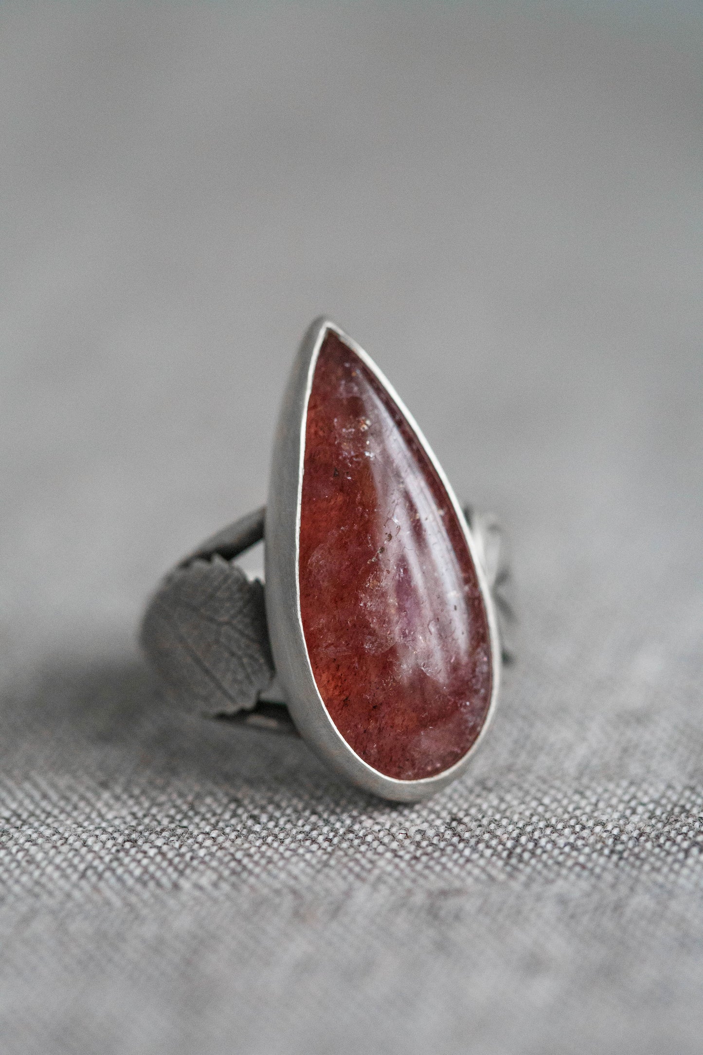 A silver ring featuring a large, teardrop-shaped reddish-brown stone, photographed close-up on a textured gray fabric background.