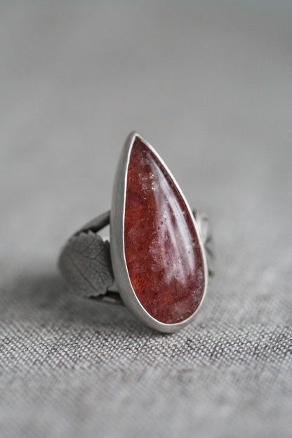 A silver ring featuring a large, teardrop-shaped reddish-brown stone, photographed close-up on a textured gray fabric background.