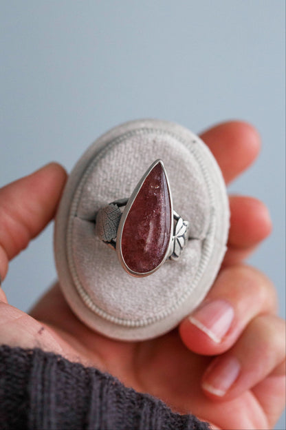 A silver ring with a teardrop-shaped reddish-brown stone, displayed on a round beige ring box held by a hand against a soft blue-gray background.
