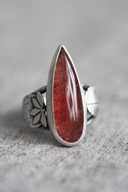 A silver ring featuring a large, teardrop-shaped red gemstone, photographed on a textured gray fabric surface with a soft-focus gray background.
