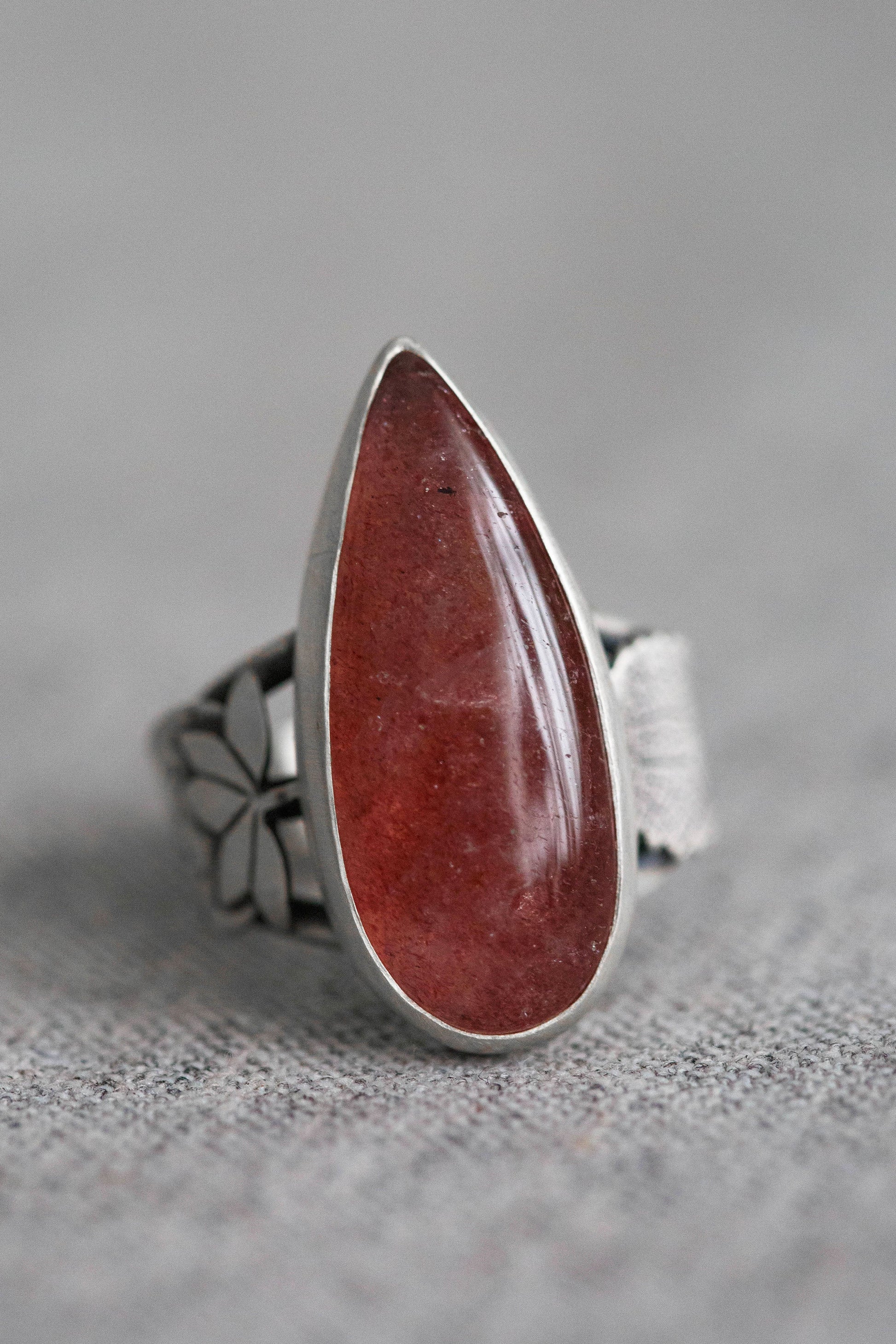A silver ring featuring a large, teardrop-shaped red gemstone, photographed on a textured gray fabric background.