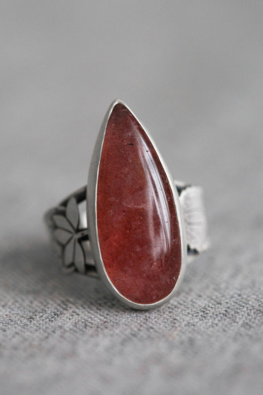 A silver ring featuring a large, teardrop-shaped red gemstone, photographed on a textured gray fabric background.
