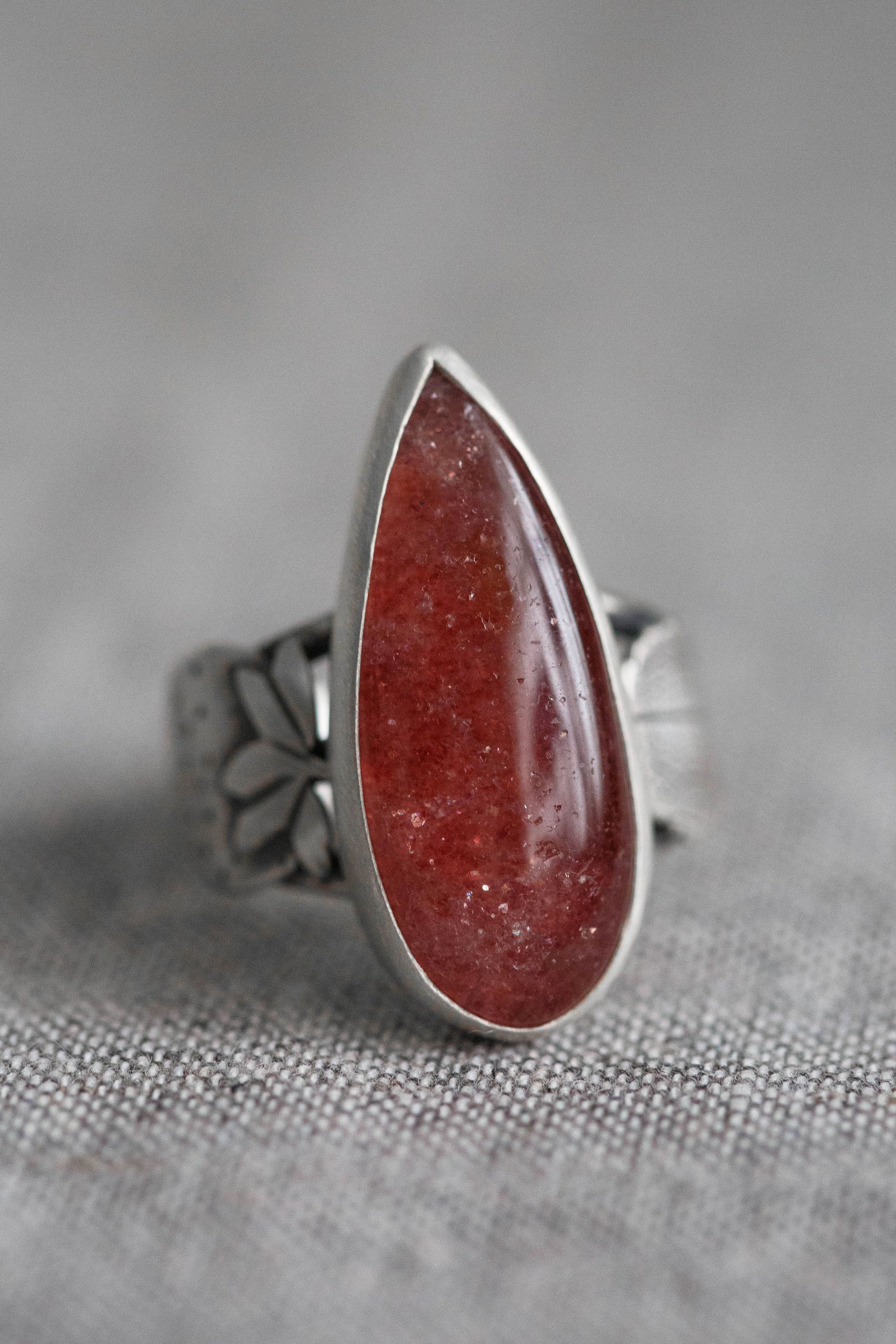 A silver ring featuring a teardrop-shaped red gemstone with tiny inclusions, set on a textured gray fabric background.