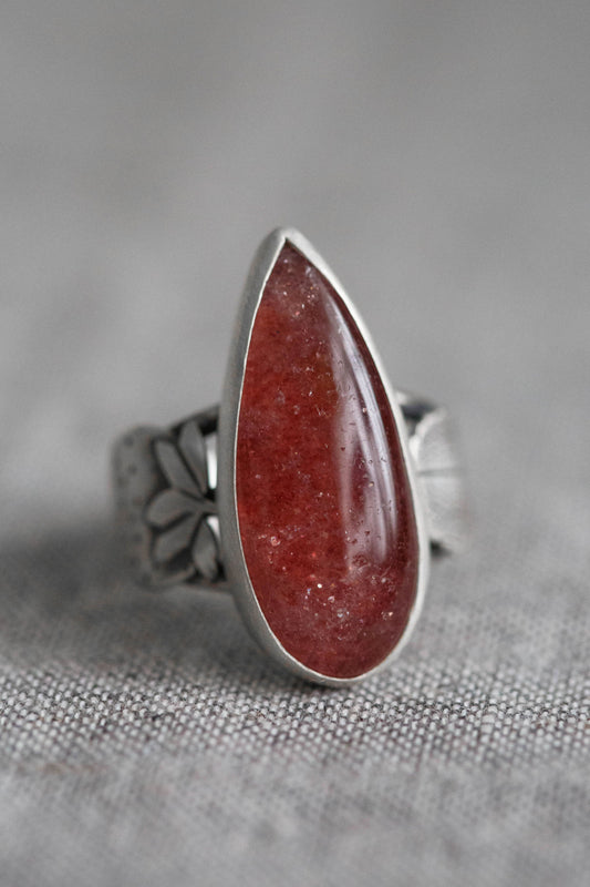 A silver ring featuring a teardrop-shaped red gemstone with tiny inclusions, set on a textured gray fabric background.