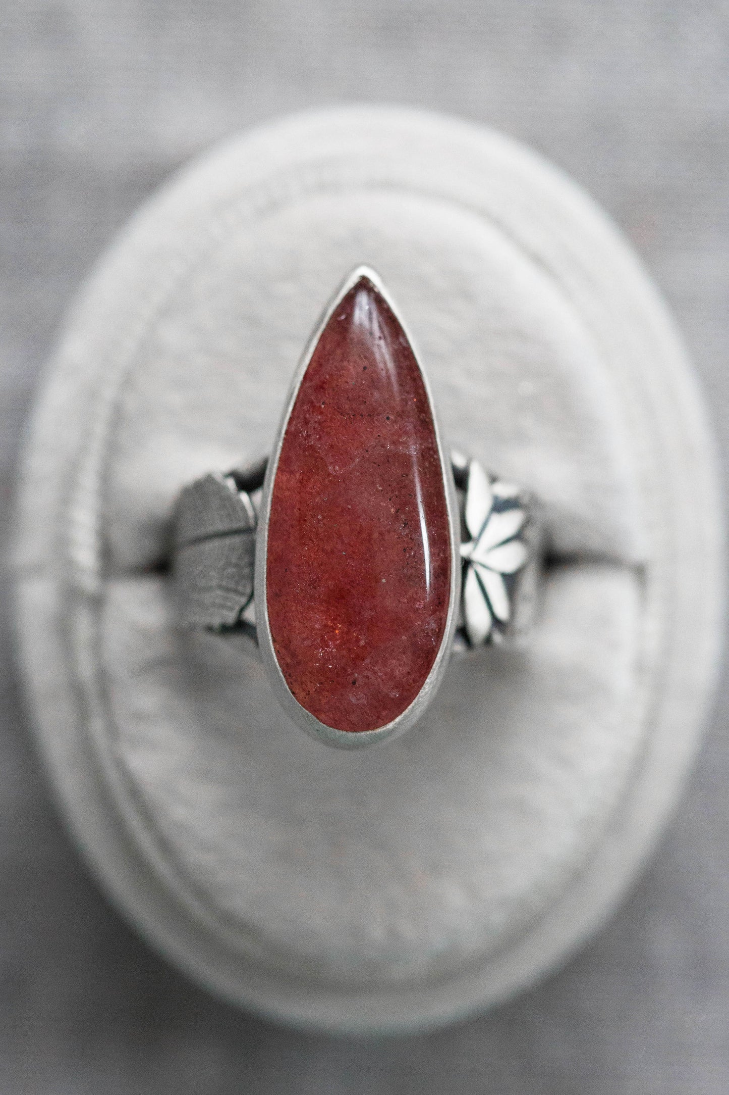 A silver ring with a teardrop-shaped red gemstone, displayed on a circular gray cushion against a textured gray background.