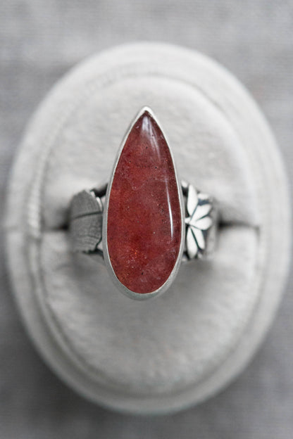 A silver ring with a teardrop-shaped red gemstone, displayed on a circular gray cushion against a textured gray background.