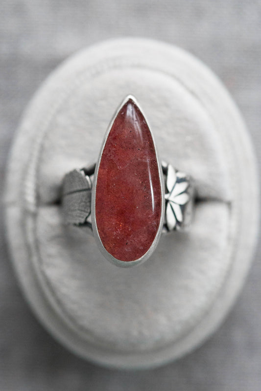 A silver ring with a teardrop-shaped red gemstone, displayed on a circular gray cushion against a textured gray background.