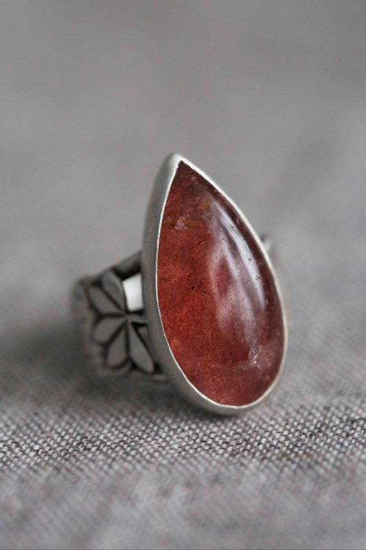 A silver ring featuring a large, teardrop-shaped reddish-brown gemstone, photographed on a textured gray fabric background.