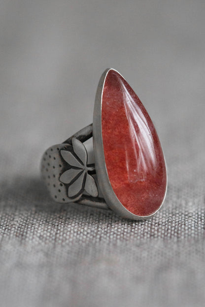 A silver ring featuring a large, polished teardrop-shaped red gemstone, set on a textured gray fabric background.