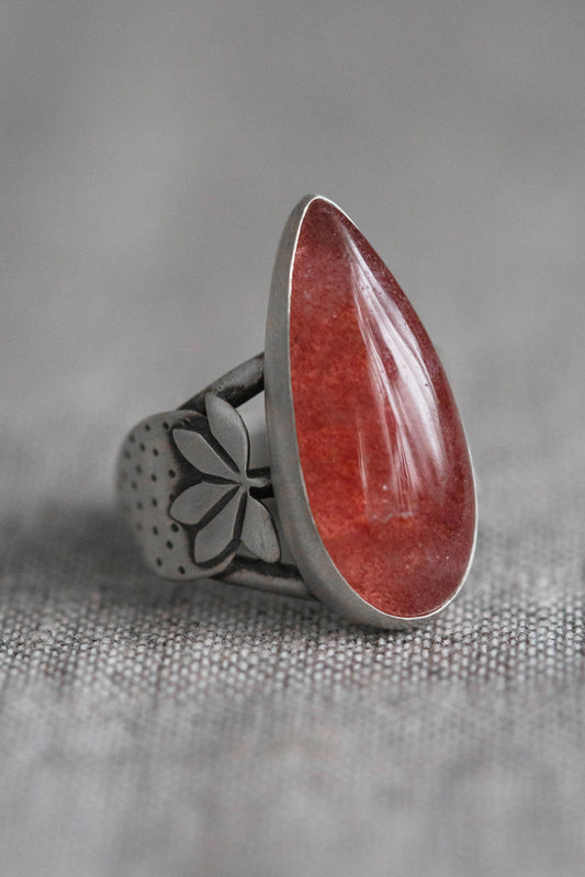 A silver ring featuring a large, polished teardrop-shaped red gemstone, set on a textured gray fabric background.