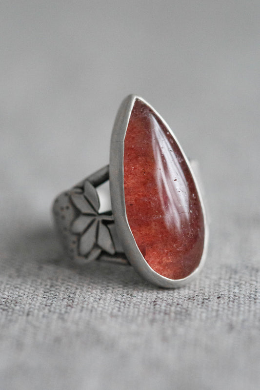 A silver ring featuring a large, teardrop-shaped red gemstone, photographed on a textured gray fabric surface.
