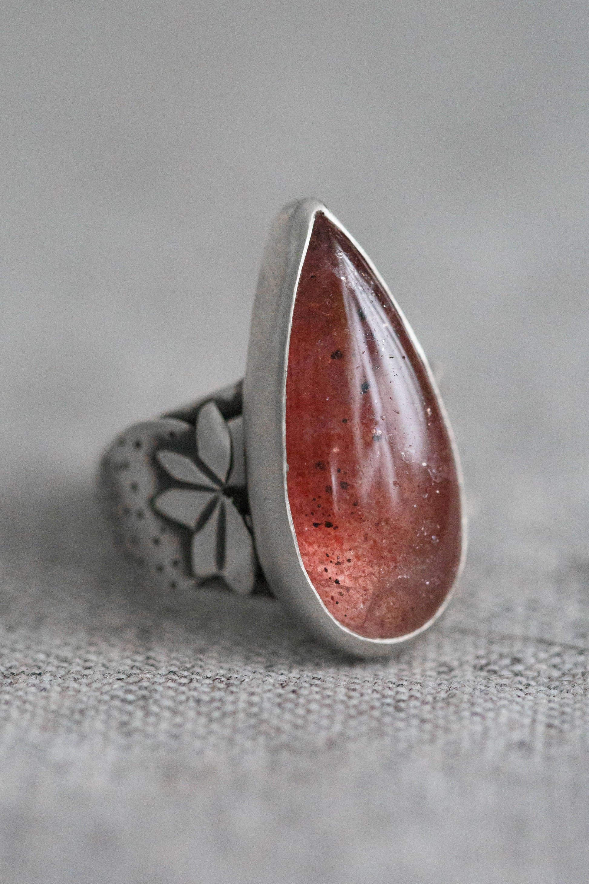 A silver ring featuring a large teardrop-shaped red gemstone with tiny gold flecks, set against a textured gray background.