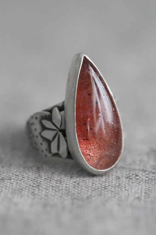 A silver ring featuring a large teardrop-shaped red gemstone with tiny gold flecks, set against a textured gray background.