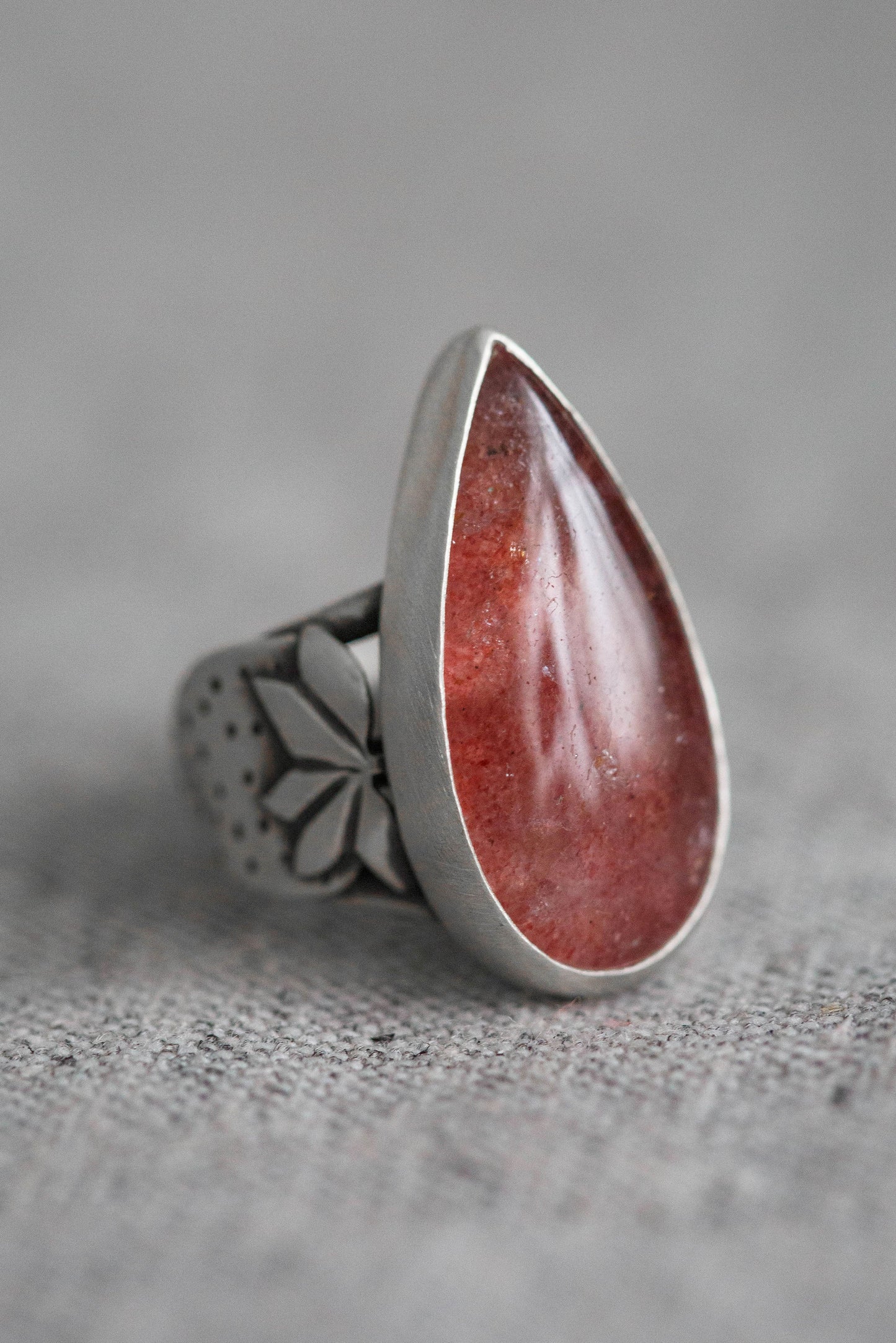 A silver ring featuring a large, teardrop-shaped red stone with a polished, glossy finish, set on a textured gray fabric background.