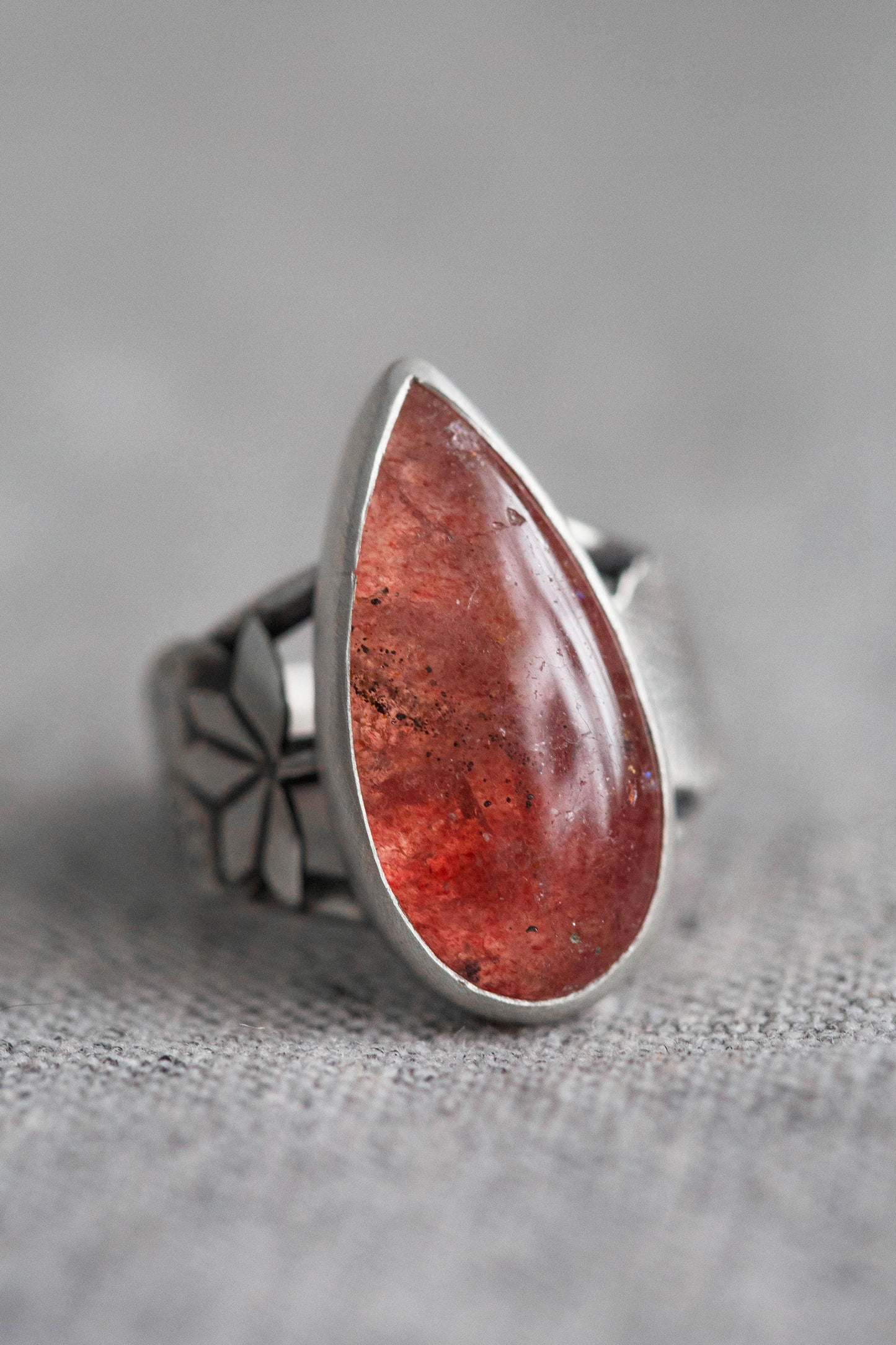 A sterling silver ring featuring a large, teardrop-shaped red gemstone with natural inclusions, set against a soft gray textured background.