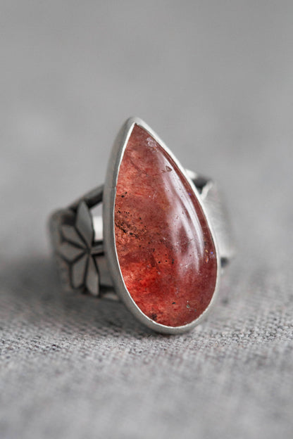 A sterling silver ring featuring a large, teardrop-shaped red gemstone with natural inclusions, set against a soft gray textured background.