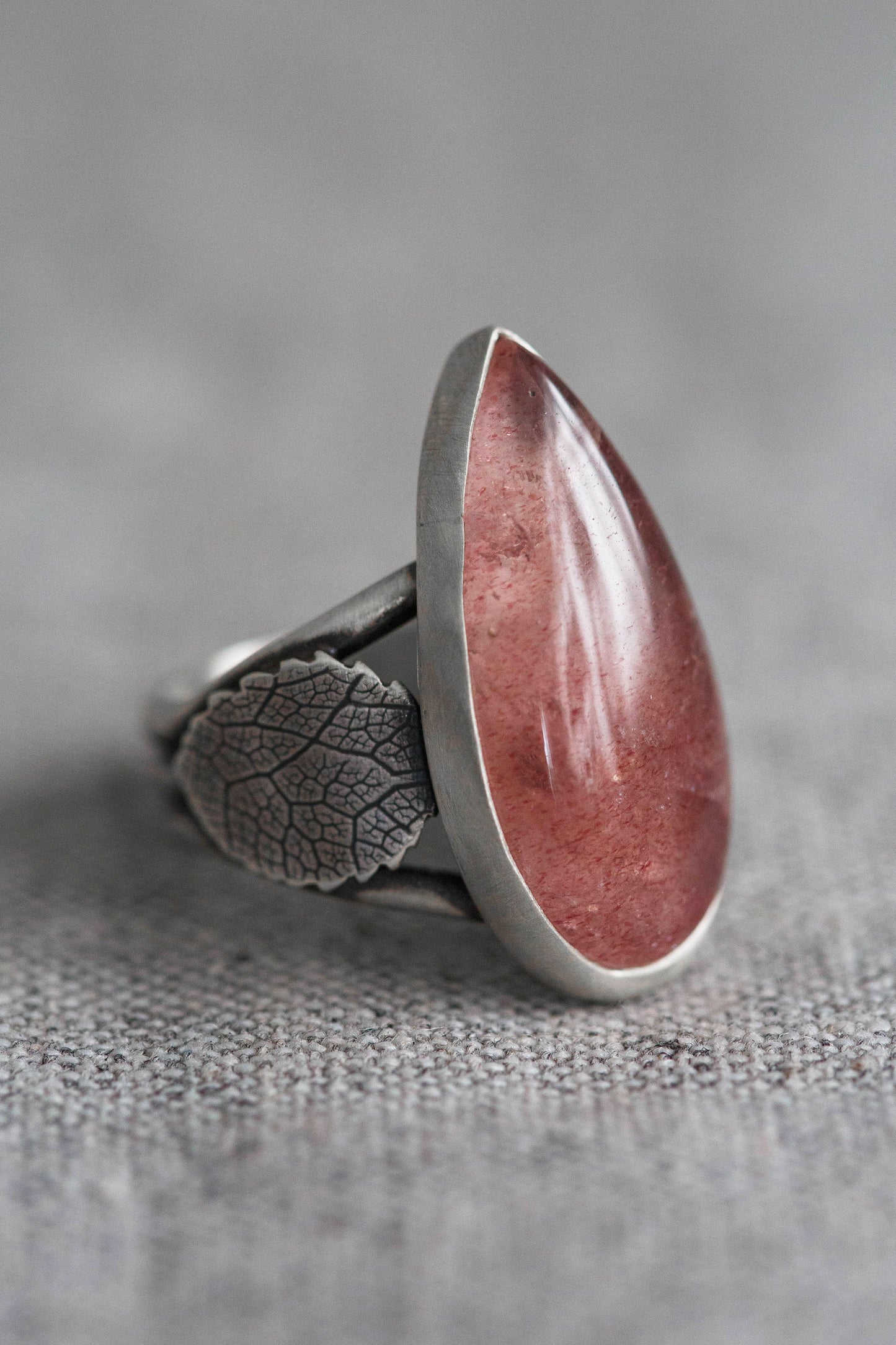A silver ring featuring a large, polished teardrop-shaped pinkish-red stone with a textured leaf engraving on the band, set against a neutral gray background.