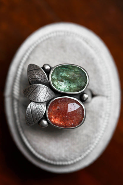 A silver ring with two oval gemstones, one green and one reddish-orange, displayed on a round white ring stand over a dark wooden surface.