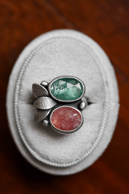 A silver ring with two oval gemstones, one green and one reddish-pink, displayed on a white textured ring box over a dark wooden surface.