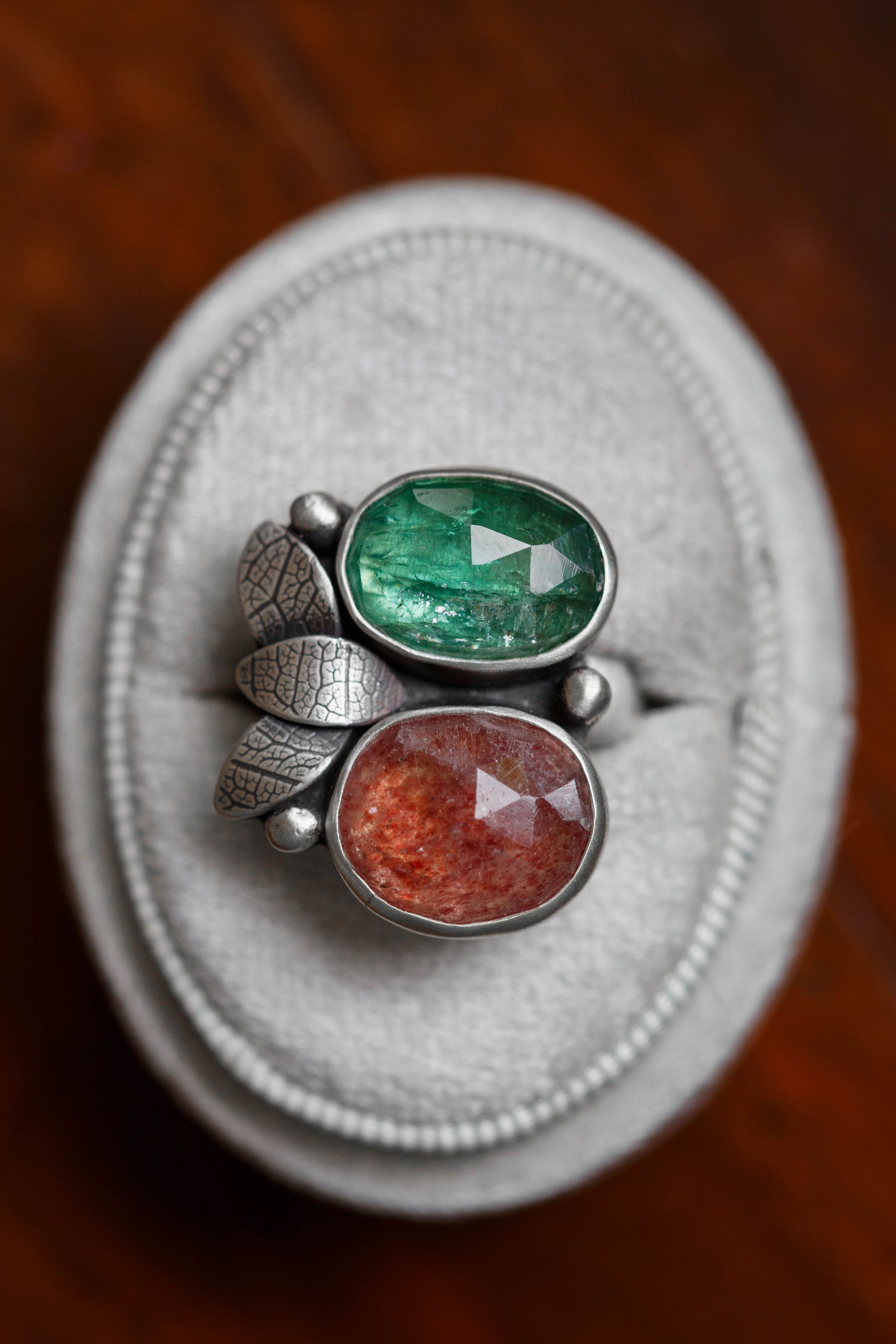 A close-up of a silver ring with two oval faceted gemstones, one green and one reddish-orange, displayed on a light gray velvet ring box over a dark wooden surface.
