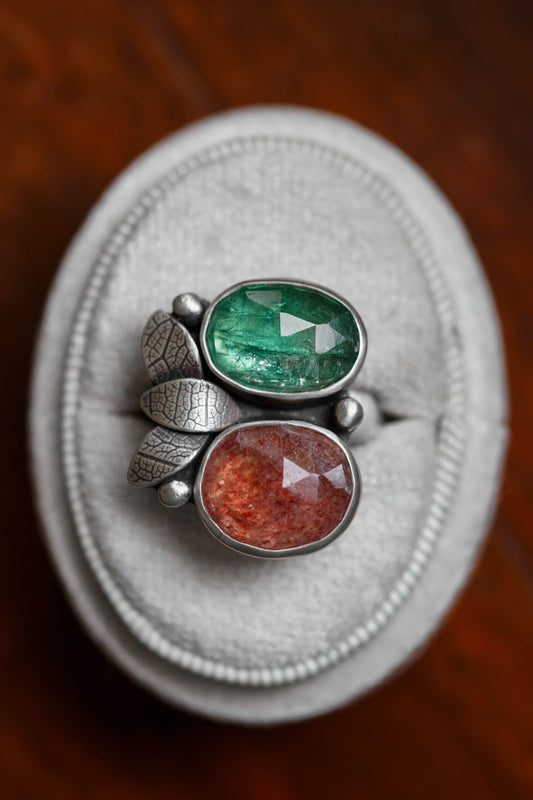 A close-up of a silver ring with two oval faceted gemstones, one green and one reddish-orange, displayed on a light gray velvet ring box over a dark wooden surface.