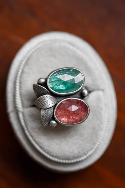 A silver ring with two oval gemstones, one green and one pinkish-red, displayed on a round gray ring stand over a dark wooden surface.