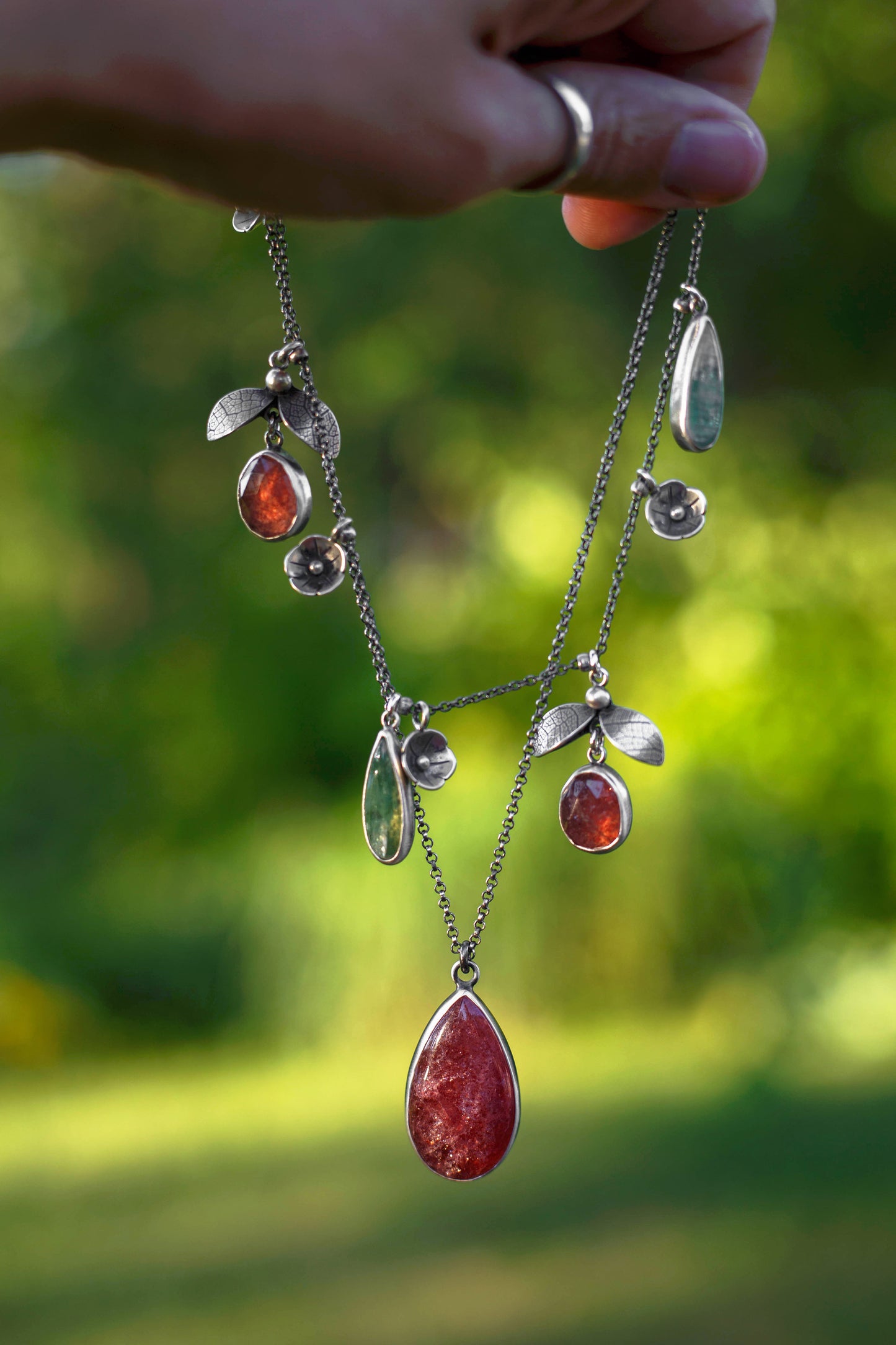 A hand holds a silver-toned leaf and teardrop pendant necklace with red and clear stones against a blurred green outdoor background.