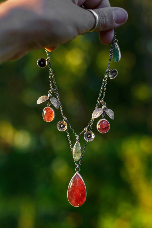 A hand holds a delicate silver-toned necklace with multiple dangling charms, including a large red teardrop pendant, small leaf charms, floral details, and colorful stones, against a blurred green outdoor background.
