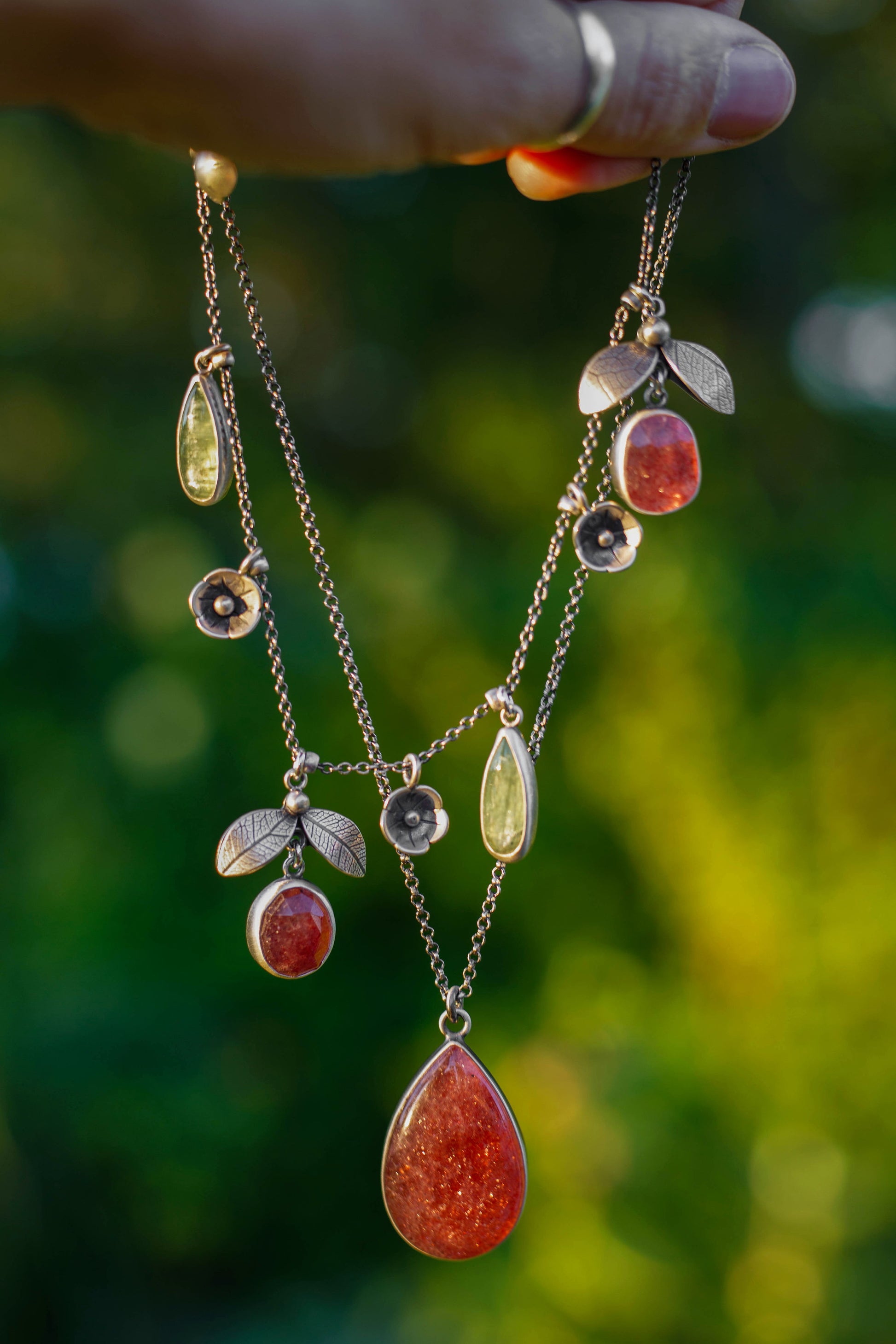 A close-up of a hand holding a layered gold necklace with teardrop and oval gemstones, including a prominent red teardrop pendant, against a blurred green outdoor background.